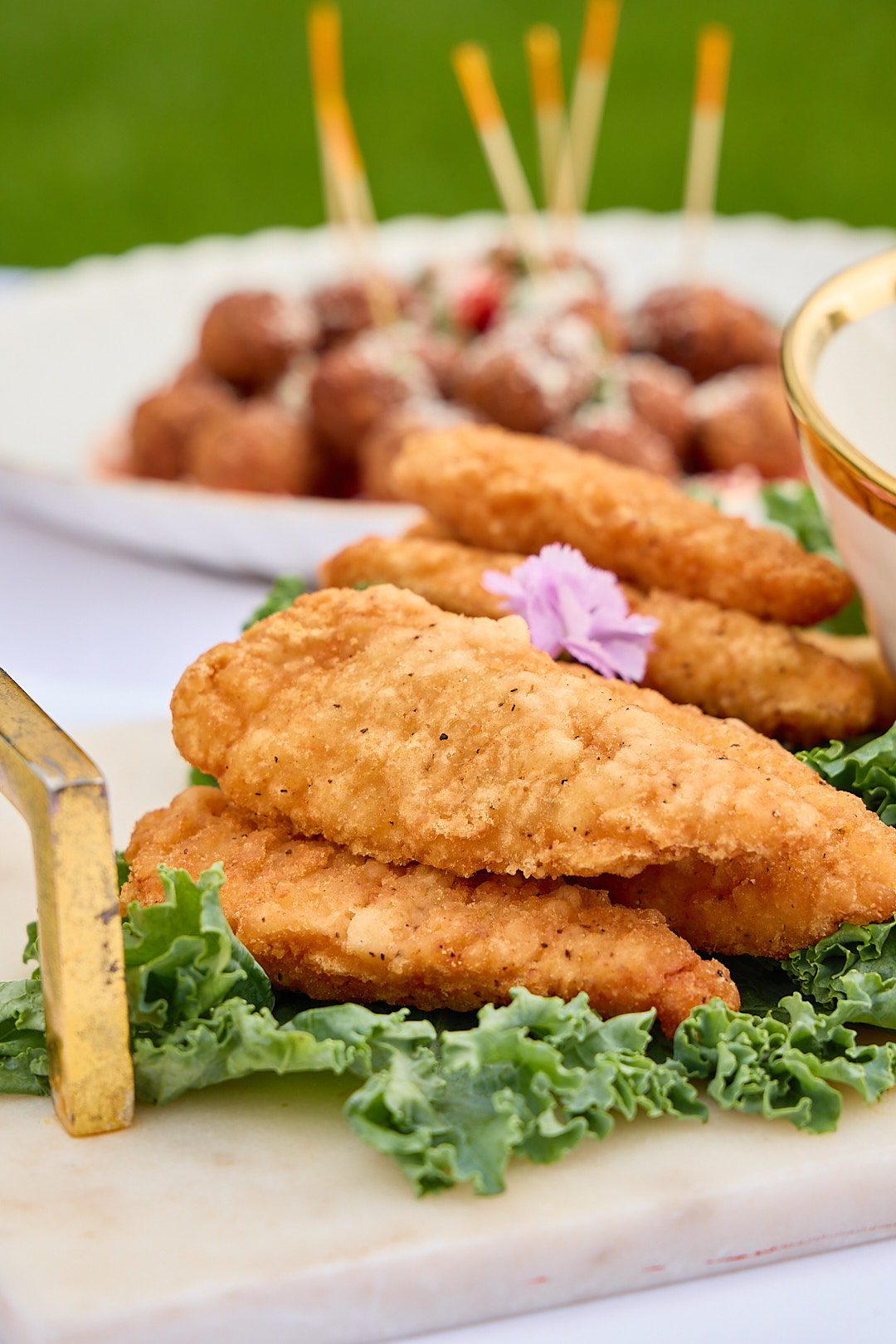 Crispy chicken tenders styled on kale with meatballs in the background.