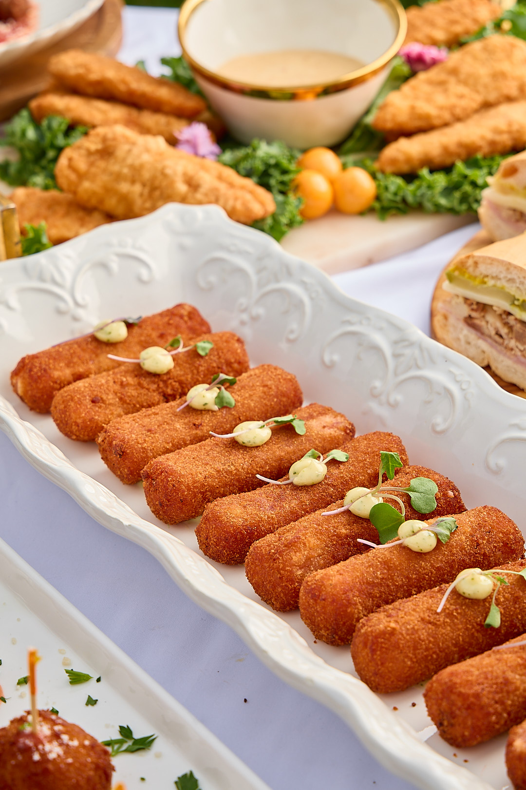 Golden Cuban croquettes on a porcelain platter with aioli, fried chicken behind.