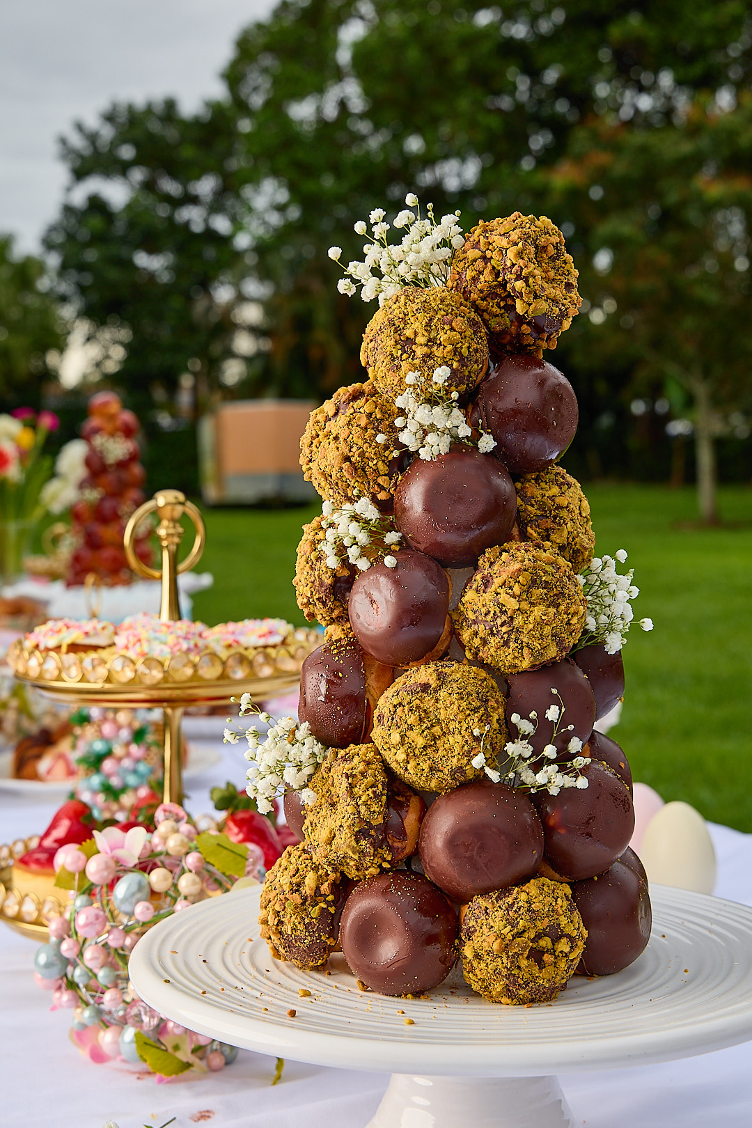 A tower of pistachio-crusted and chocolate-glazed profiteroles on a cake stand at golden hour.
