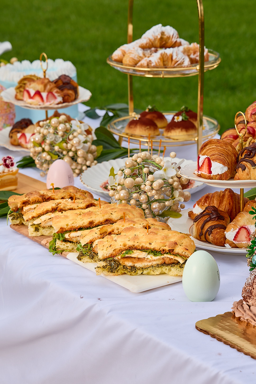 A spring catering setup with focaccia sandwiches, croissants, brioche, and floral arrangements on white linen.