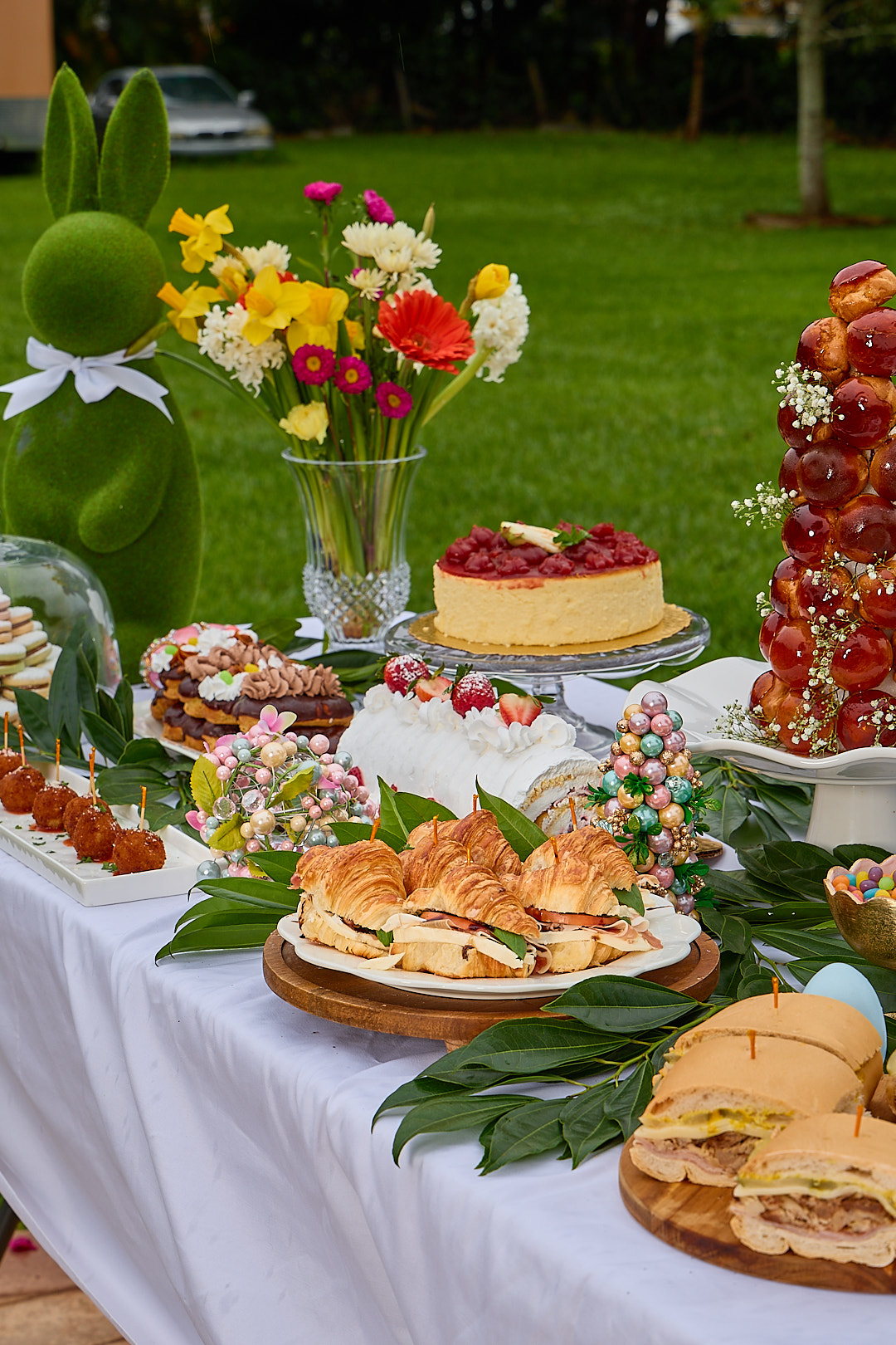A wide spring catering tablescape: cheesecake with strawberries, prosciutto croissants, brioche tower, croquette skewers.